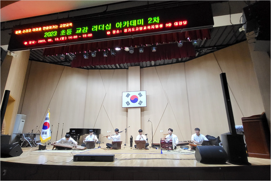 전통음악집단 샛의 공연 사진이다. 강당 무대 위에서 6명의 단원이 나란히 앉아 제각각 전통 악기를 연주하고 있다. 무대 중간과 왼쪽엔 태극기가 있다.